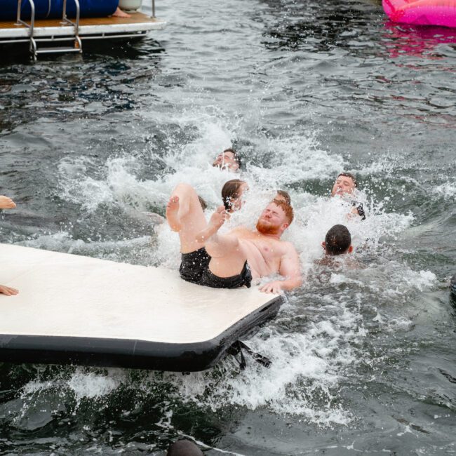 A group of people swim in a lake near boats and a large inflatable flamingo. A man is in mid-fall from a floating platform, creating a splash. Other swimmers surround him, with one person looking up at him and another reaching out. Perfect for boat rental and parties with The Yacht Social Club Sydney Boat Hire.