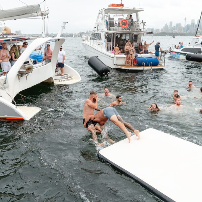 A group of people enjoy a lively time in the water between two boats, some swimming while others socialize on deck. Two men playfully wrestle on a floating mat. In the background, more anchored boats and a cityscape with high-rise buildings set the scene for The Yacht Social Club Sydney Boat Hire.