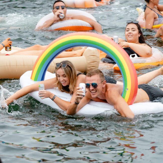 A group of people enjoying a sunny day on a lake, floating on various inflatable rafts. Two individuals in the foreground are on an inflatable with a rainbow arch, smiling and holding drinks, while others in the background relax on different floaties. It's like The Yacht Social Club Sydney Boat Hire fun on water.