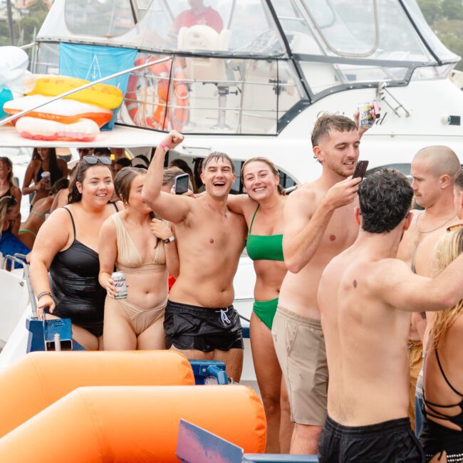 A large group of people in swimsuits are having a lively party on a boat. Some are dancing, others are holding drinks, and everyone appears to be in high spirits. Inflatable pool toys and a slide are visible, indicating a fun, water-themed gathering hosted by The Yacht Social Club Event Boat Charters.