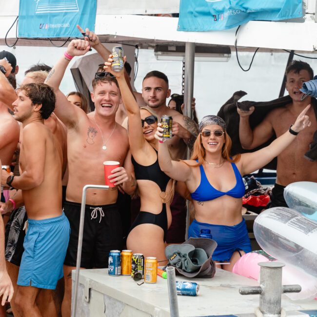 A group of people in swimwear are celebrating on a boat, holding drinks and smiling at the camera. Some are wearing sunglasses, enjoying the party atmosphere. Inflatable floats and drink cans are visible around them. This scene captures the fun of The Yacht Social Club Event Boat Charters in Sydney.