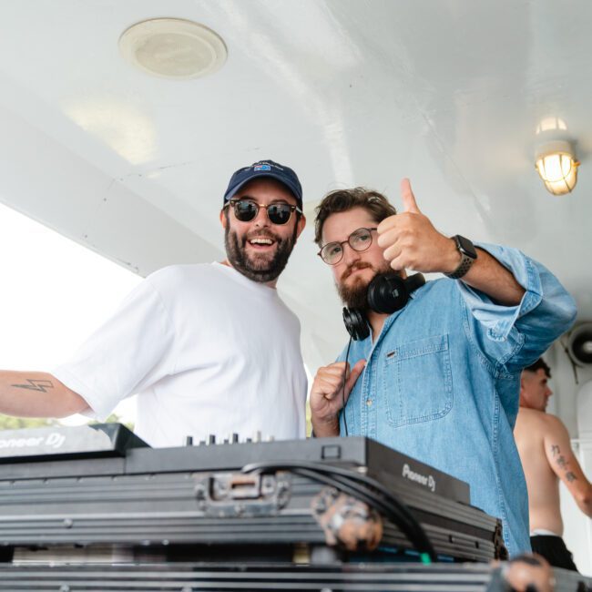 Two men are posing happily in a boat's interior at The Yacht Social Club Sydney Boat Hire. One is dressed in a white T-shirt and blue cap, flashing a peace sign and wearing sunglasses. The other, wearing glasses and a denim shirt, gives a thumbs-up. DJ equipment is in the foreground.