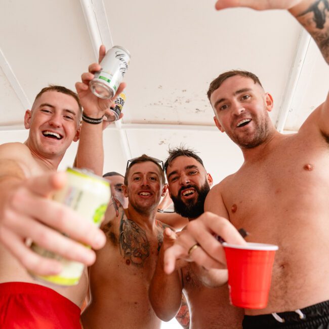 A group of shirtless men at a party on a yacht, holding cans of drinks and red cups, smiling and gesturing enthusiastically at the camera. Some of them have tattoos. They appear to be enjoying a lively and festive atmosphere hosted by The Yacht Social Club Sydney Boat Hire.