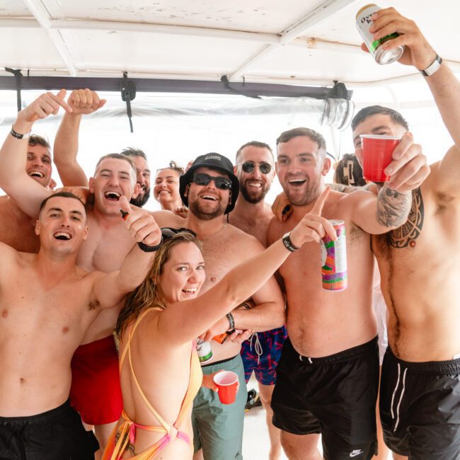 A joyful group of friends in swimwear poses together on a boat, all smiling and raising drinks. They appear to be celebrating, enjoying the sunny weather, and having a great time. The background shows a sunny day with partly cloudy skies—courtesy of Luxury Yacht Rentals Sydney.