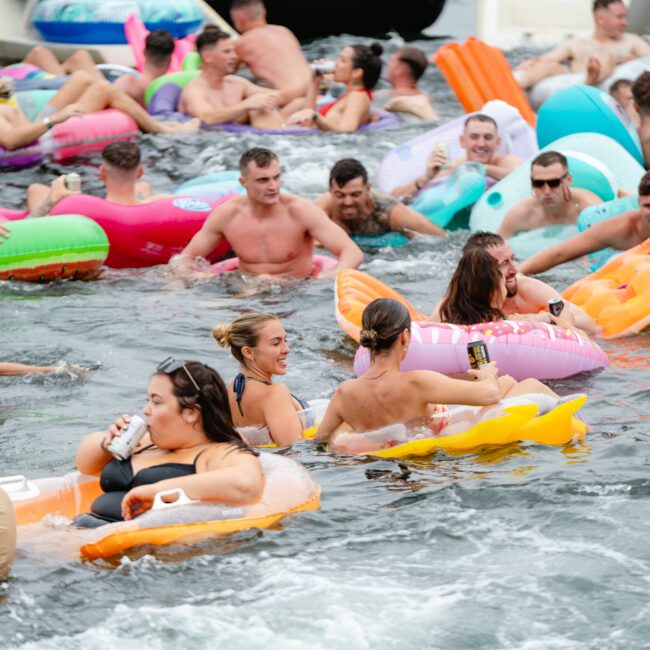 A large group of people enjoying a sunny day on the water with various colorful inflatable floats. Some are drinking from cans and bottles, while others chat and relax. The atmosphere is lively and cheerful, with everyone having a good time, reminiscent of boat parties by The Yacht Social Club in Sydney Harbour.