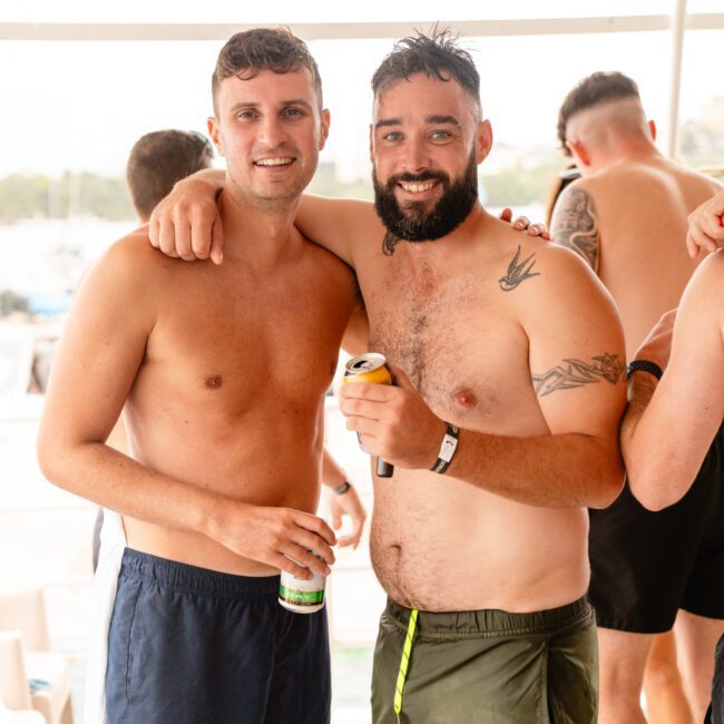 Two men in swim trunks are standing side by side, smiling at the camera aboard a luxury yacht. The man on the left has short hair and is wearing navy blue trunks, while the bearded man on the right, with tattoos on his arm, is wearing olive green trunks and holding a drink. Other people are in the background.