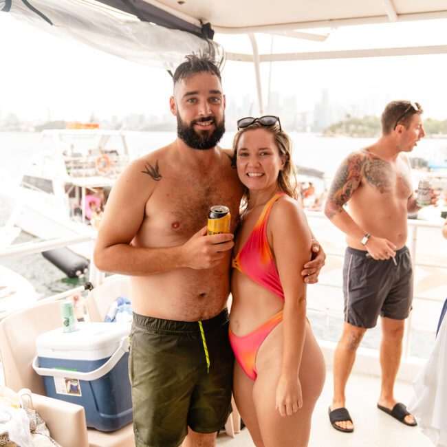 A bearded man and a woman in a pink swimsuit smile and pose together on a boat rental from The Yacht Social Club. The man holds a canned drink. Other people are in the background, with boats and Sydney's cityscape visible in the distance. The atmosphere is relaxed and sunny, perfect for Boat Parties Sydney.
