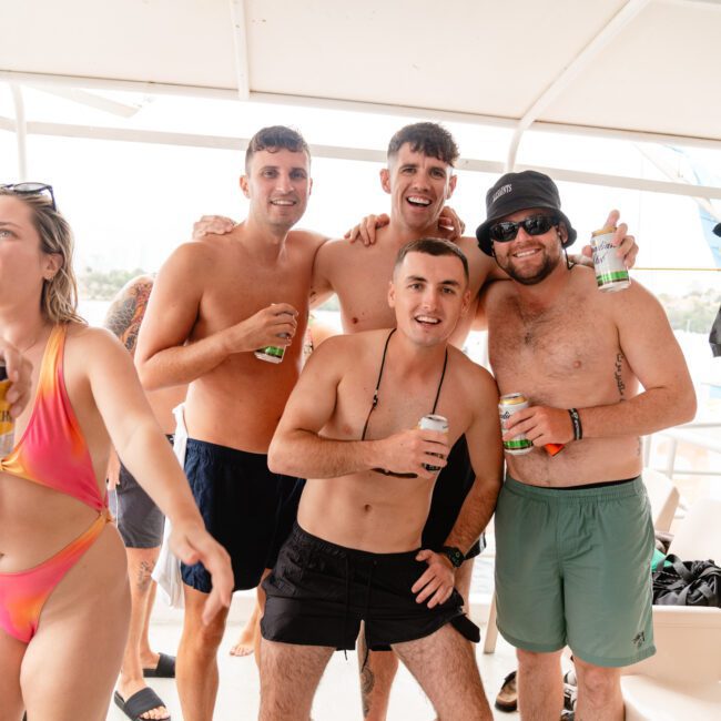Four men in swimwear, smiling and holding drinks, stand together under a boat canopy. One man wears a hat and sunglasses. A woman in a colorful bikini walks by on the left side of the image. The group appears relaxed, enjoying their time at The Yacht Social Club Event Boat Charters.