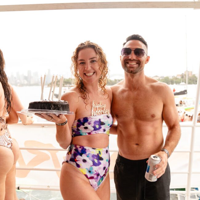 Two people are smiling on a boat. The person on the left is holding a small cake with candles and wearing a multicolored swimsuit. The person on the right is shirtless, wearing black shorts, and holding a can. They appear to be enjoying a sunny day by the water, likely part of Sydney Harbour Boat Hire The Yacht Social Club.
