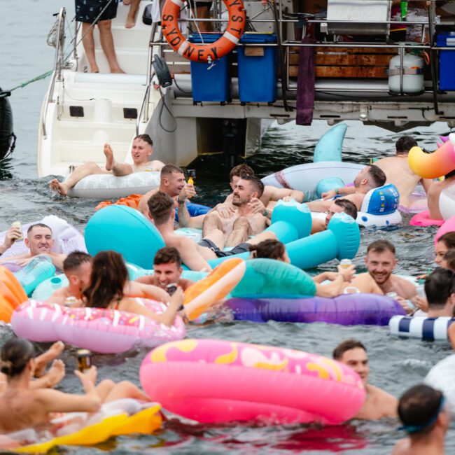 A lively group of people in colorful inflatable pool floats enjoy a party in the water near a boat. Some floats are shaped like animals and objects. The anchored boat, part of The Yacht Social Club Sydney Boat Hire, is equipped with life rings, and more people are on the deck. The scene is festive and energetic.