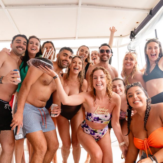 A group of people celebrate on a luxury yacht. A woman in the center holds a birthday cake with lit candles. Everyone around her is smiling, with some raising their hands in joy. Most are dressed in swimwear, indicating a festive, aquatic outing with Sydney Harbour Boat Hire The Yacht Social Club.