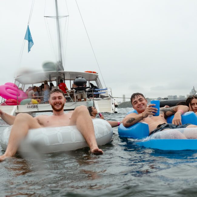 Two men are lounging on inflatable floats in the water, smiling and holding drinks. Behind them are two women on another float, and a yacht in the background. The atmosphere is relaxed and joyful, suggesting a fun day with The Yacht Social Club Event Boat Charters.