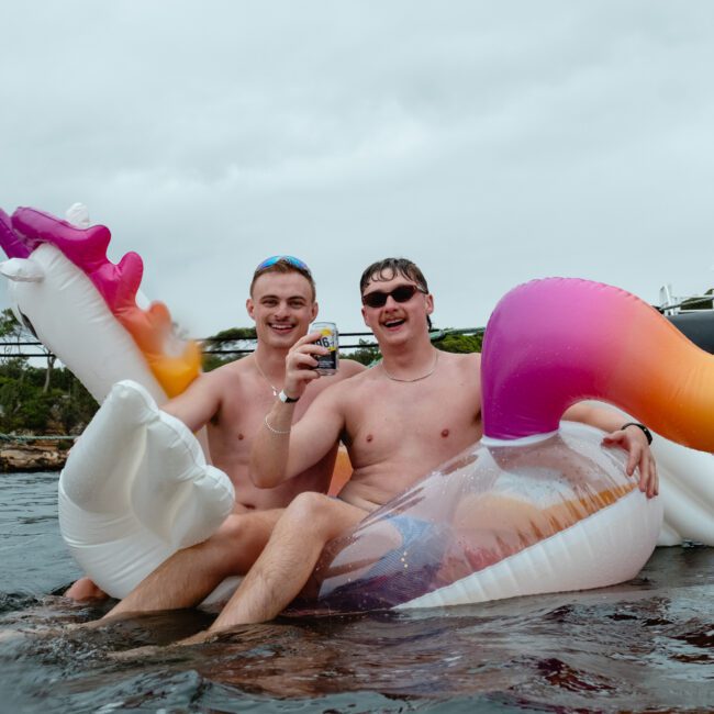 Two shirtless men are sitting on a colorful unicorn float in the water, smiling and holding drinks during one of the Boat Parties Sydney The Yacht Social Club events. They are surrounded by water with boats in the background and appear to be enjoying a relaxing time outdoors.