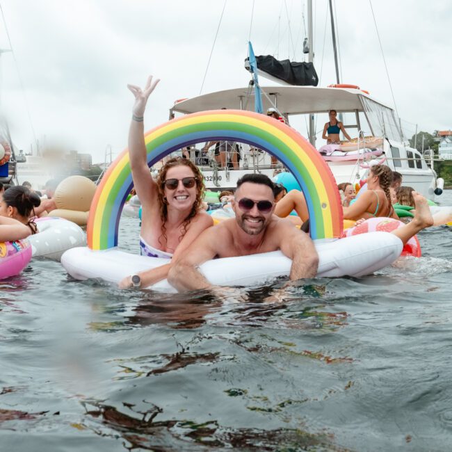 A man and a woman are floating on a large inflatable pool float shaped like a rainbow in a crowded water area. Other people on colorful inflatables, boats, and yachts are visible in the background. The woman is smiling and holding up a peace sign. It's an ideal day for Boat Rental and Parties Sydney The Yacht Social Club.