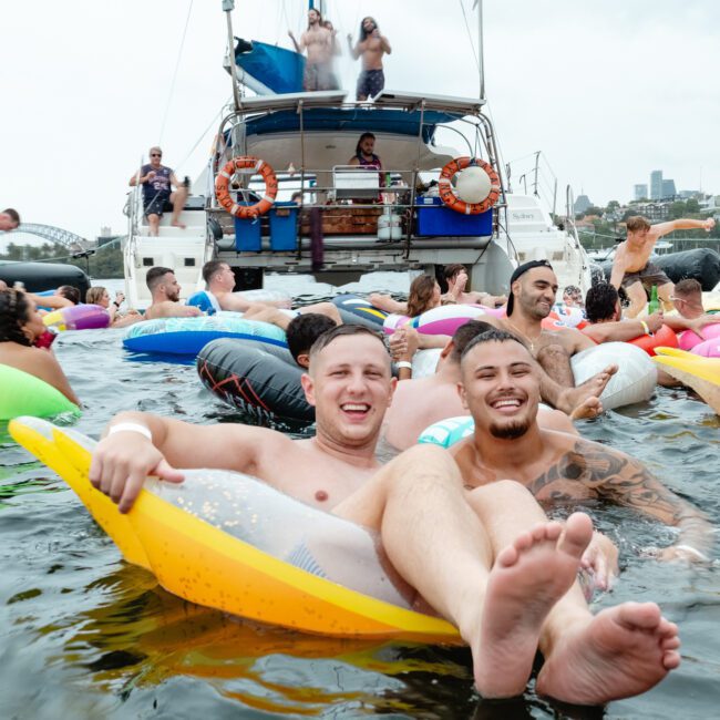 A group of people enjoying a summer party on the water, with two men smiling and floating on inflatable toys in the foreground. Others are swimming or lounging on inflatables nearby, with boats and city buildings visible in the background—a perfect scene from The Yacht Social Club Sydney Boat Hire.