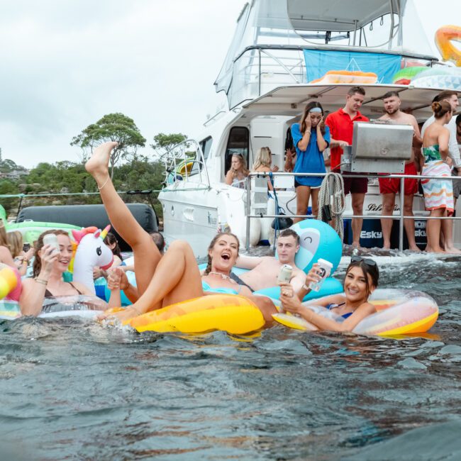 A lively boat party hosted by The Yacht Social Club with people enjoying themselves on inflatable floats in the water. The boat's deck is crowded with socializing guests, while others float on colorful inflatables, including a unicorn and donut. Lush greenery is visible in the background.