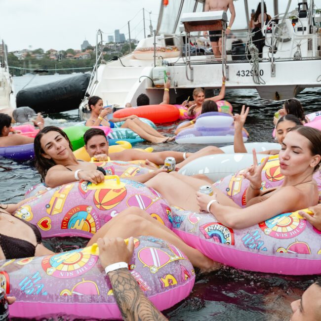 A group of people enjoying a party in the water, lounging on colorful inflatable tubes and sipping drinks. They are near several docked boats, creating a lively and festive atmosphere with The Yacht Social Club Sydney Boat Hire. Some people are making peace signs and smiling at the camera.