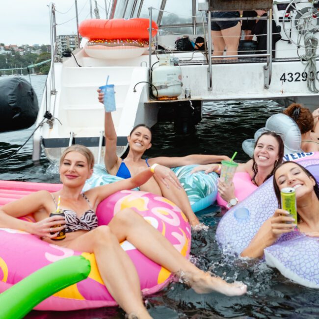 A group of four women wearing swimsuits are relaxing on colorful inflatable rings beside a boat. Smiling and holding drinks, they seem to be enjoying a sunny day out with Sydney Harbour Boat Hire The Yacht Social Club. Other boats and a city skyline can be seen in the background.
