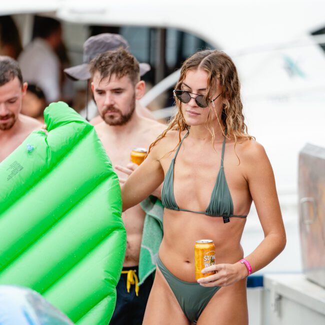 A woman in a green bikini and sunglasses holds a green inflatable float and a canned beverage. She is surrounded by other people, including a bearded man with a towel and drink. In the background, boats from The Yacht Social Club are docked, hinting at the vibrant boat parties on Sydney Harbour.
