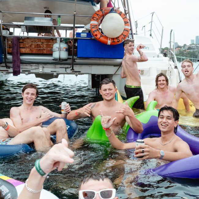 A group of seven young men are enjoying a day on the water, lounging on inflatables with drinks in hand and smiles on their faces. They are gathered near The Yacht Social Club, everyone is in swimwear, enjoying a casual, fun-filled atmosphere typical of Sydney Harbour Boat Hire events.