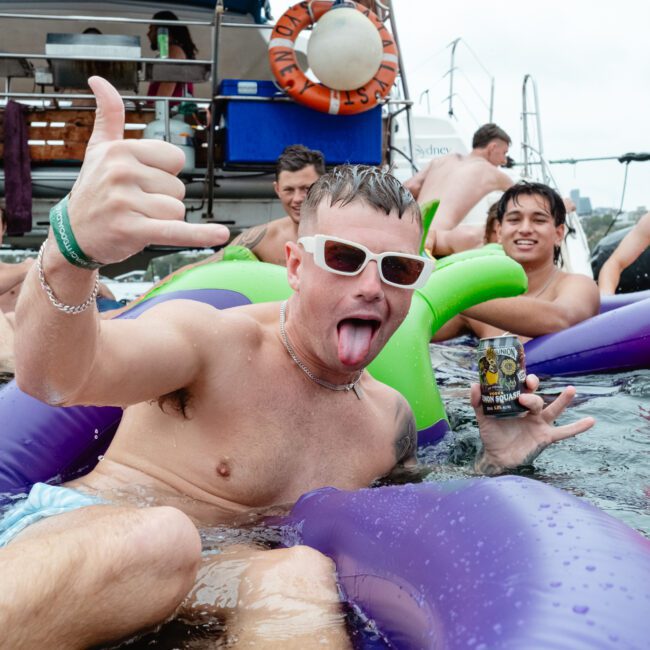 A group of young men are having fun in a pool on inflatable floats. One man in the foreground sticks out his tongue, makes a hand gesture, and holds a beer can. He wears sunglasses and bracelets. Others in the background are smiling and enjoying the water, reminiscent of The Yacht Social Club's vibrant boat parties in Sydney Harbour.