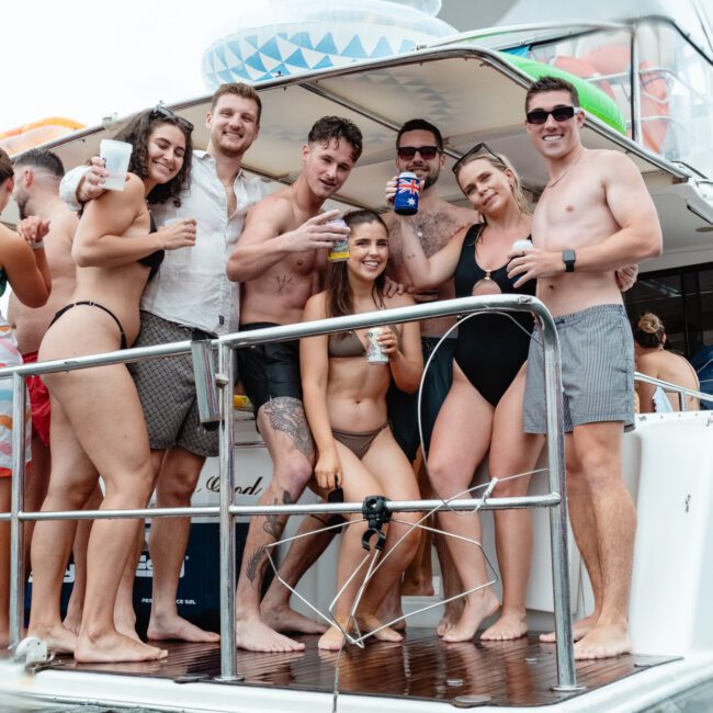A group of people in swimsuits are standing on the deck of a boat, smiling and holding drinks. The Yacht Social Club Sydney Boat Hire is partially visible on the water, suggesting a fun day out. Some are standing close together, and others are seated, with a festive atmosphere evident.