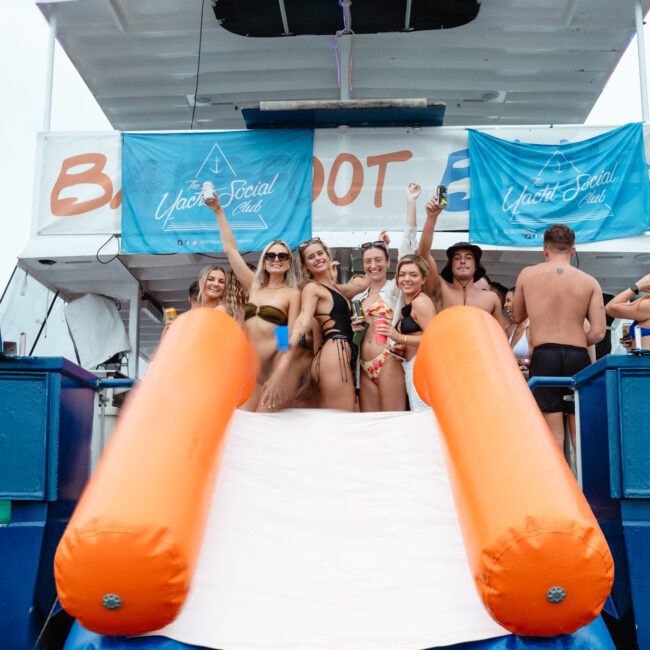 A group of people in swimwear are on a boat with blue banners reading "Yacht Social Club." The boat has an orange-and-white slide at the end, leading into the water. Some people are posing and smiling for the camera, while others enjoy the background during this Boat Parties Sydney The Yacht Social Club event.