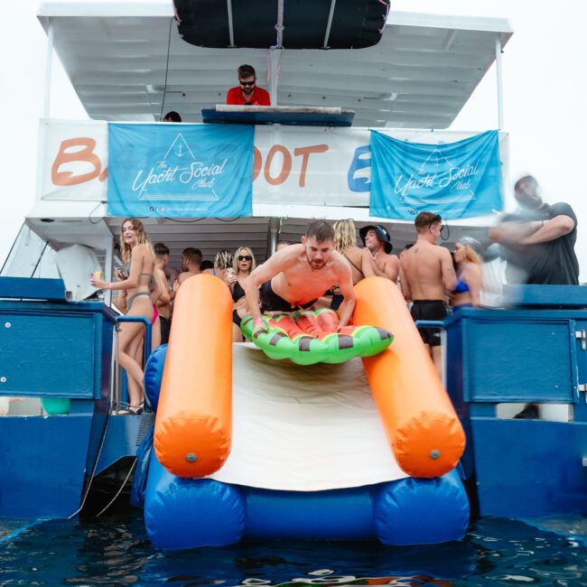 A group of people enjoys a party on a boat. One person slides down an inflatable water slide into the sea, holding a fruit-shaped float. Others watch and cheer, with blue banners reading "White Sails" visible on the luxury yacht rental from Sydney Harbour Boat Hire The Yacht Social Club.