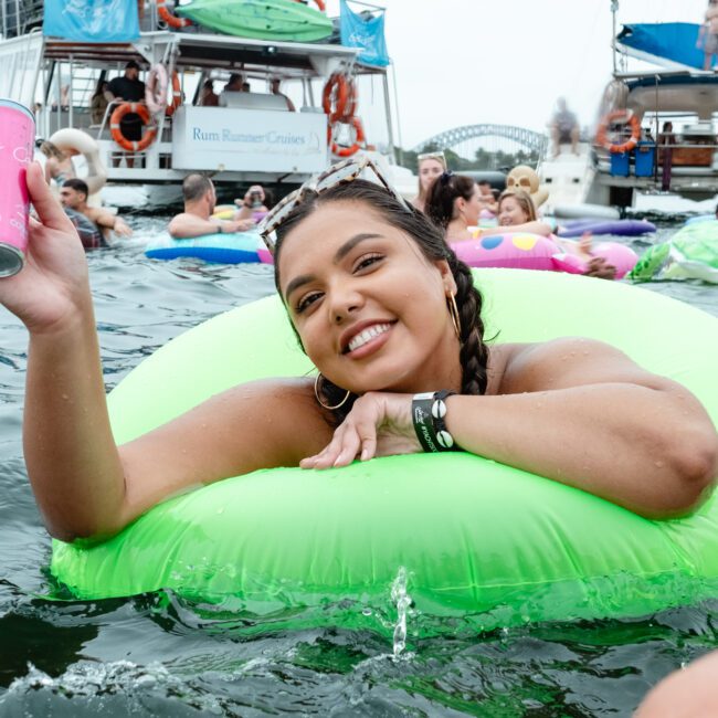 A woman smiling and holding a can of drink is lounging in a green pool float in the water. Behind her, people are enjoying themselves on various inflatables and boats, with a cityscape and bridge visible in the background. The scene is lively and festive, showcasing the vibe of Sydney Harbour Boat Hire at The Yacht Social Club.