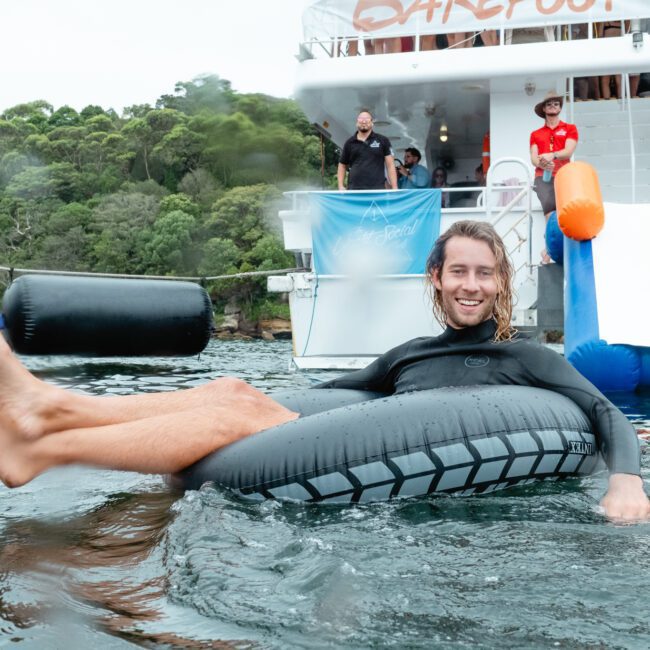 A man wearing a black wetsuit is floating on an inner tube in the water near a boat with a slide. He has long hair and is smiling. Other people are on the boat, enjoying The Yacht Social Club Sydney Boat Hire experience, and there's a forested area in the background.