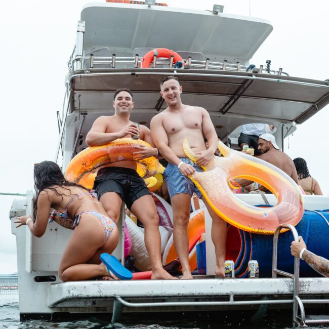 Two shirtless men stand on the back of a boat holding large inflatable pool toys, smiling towards the camera. A woman in a colorful swimsuit kneels near them on the boat, while another person holds onto the boat from the water. More people are on the Luxury Yacht Rentals Sydney vessel in the background.