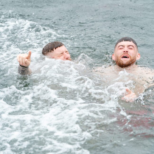 Two men swimming in open water. One is gesturing with a raised index finger while both wear expressions of exertion. They appear to be splashing through waves, suggesting they are engaged in a challenging activity or competition, perhaps near where The Yacht Social Club Event Boat Charters take place.
