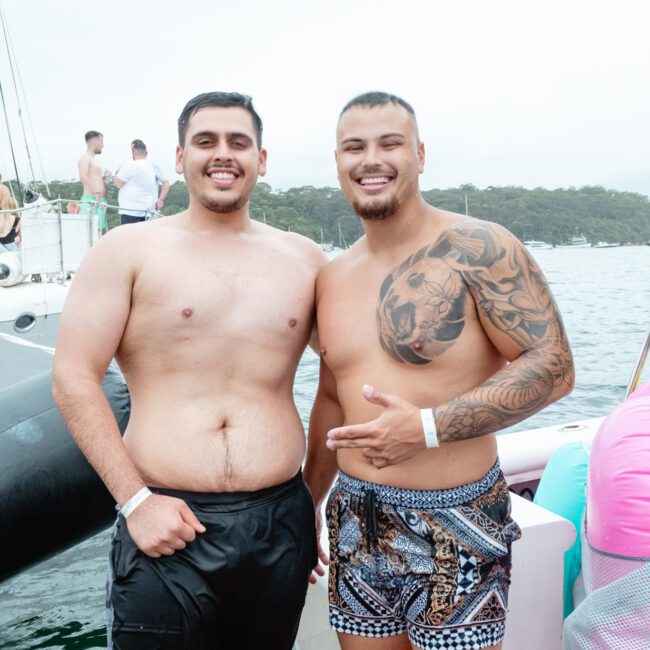 Two smiling men with short dark hair pose shirtless on a boat. The man on the right, sporting a tattoo, points at the other man. Both wear swim shorts against a scenic backdrop of water and trees. Experience this with The Yacht Social Club Sydney Boat Hire for your next adventure.