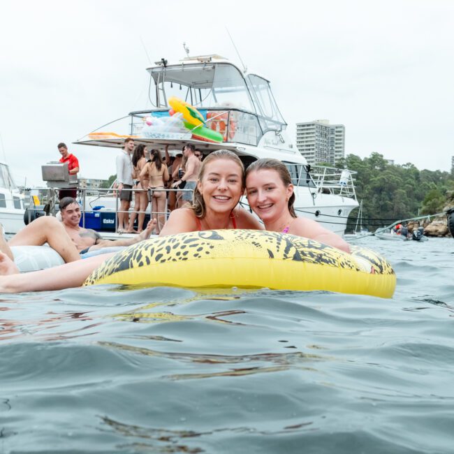 Two women float on a leopard-print inflatable in a body of water. People enjoy themselves on nearby inflatables and boats, hinting at The Yacht Social Club event. Trees and buildings dot the distance, suggesting a lively outdoor gathering near the shoreline.
