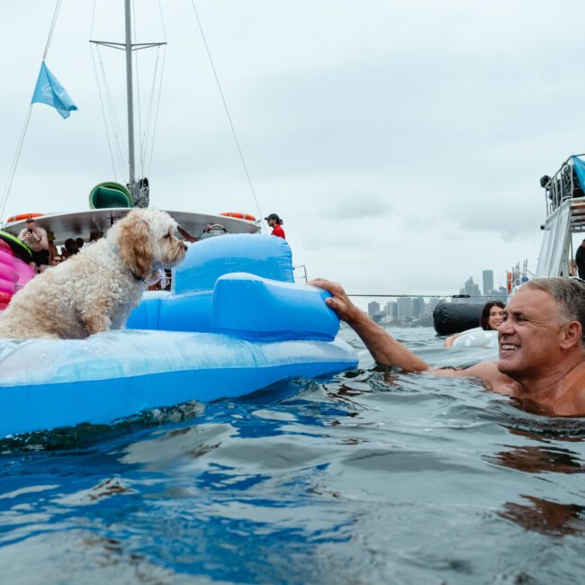 People enjoying a day on the water; a man swims towards a fluffy white dog sitting on a blue inflatable raft. Other people on inflatables and boats are visible in the background. The sky is overcast, and city buildings can be seen in the distance, creating an ideal setting for Sydney Harbour Boat Hire The Yacht Social Club.