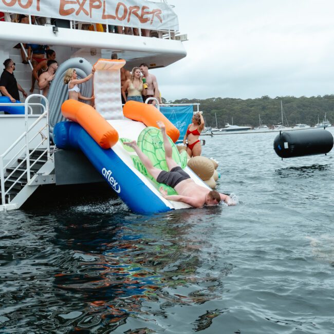 A group of people is enjoying a boating adventure with The Yacht Social Club. Some are on the boat while others are sliding down an inflatable slide into the water. A person is halfway down the slide, and another is swimming nearby. Other boats and a cloudy sky are in the background.