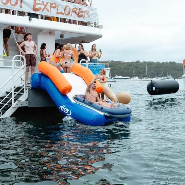 A group of people are enjoying a boat party hosted by The Yacht Social Club. Some are standing on the deck while others are riding down an inflatable slide into the water. Other boats are visible in the background, and the scene is lively with people in swimsuits having fun.