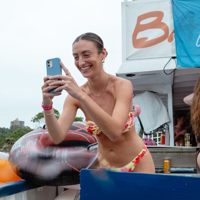 A woman in a bikini smiles and holds up her phone, taking a photo or video. She is on a yacht at The Yacht Social Club Sydney Boat Hire with others, some of whom are also holding drinks. The background shows part of the boat with banners and a blurred shoreline with trees and buildings.
