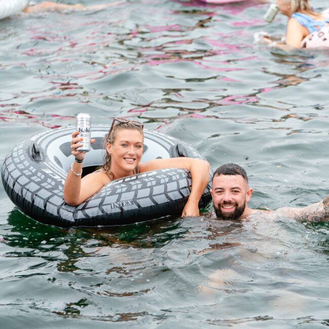 A woman in a grey inflatable ring holds up a beverage can while smiling at The Yacht Social Club event. A man, partially submerged in water, grins next to her. People on colorful inflatables are seen enjoying the water in the background.