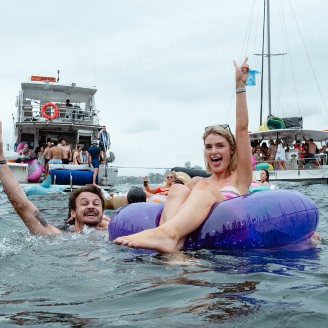 Several people enjoy a day out on the water with inflatable rafts and floaties. The image captures a smiling man splashing in the water next to a woman sitting on a purple float, raising her hand in a joyful gesture. Boats and people from Sydney Harbour Boat Hire The Yacht Social Club create a festive atmosphere.