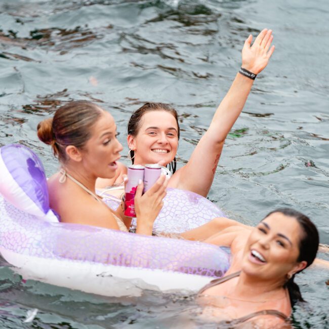 Three people are enjoying themselves in the water. One person lounges on an inflatable ring holding a drink, another waves at the camera, and a third person smiles while swimming. A pink and white inflatable ball floats nearby, setting the perfect scene for The Yacht Social Club Sydney Boat Hire experience.