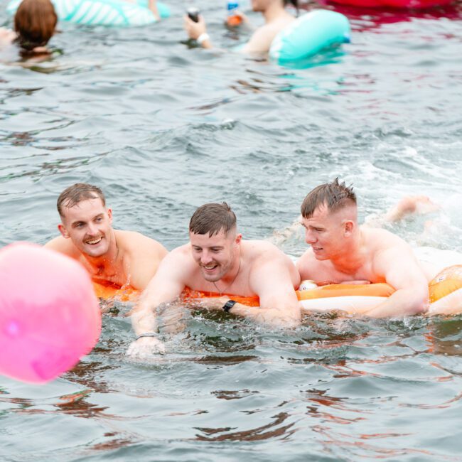 Three men are floating on an inflatable float in a body of water, enjoying themselves. One of them holds onto the edge of the float while a pink balloon drifts nearby. Other floats and swimmers can be seen in the background, making it feel like a Boat Parties Sydney The Yacht Social Club scene.