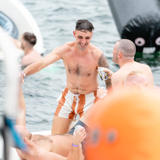 A group of men enjoy a sunny day on the water. One man in orange and white swim trunks stands smiling, holding a can, while others relax on floating inflatables. They appear to be having a lively and fun time outdoors, perhaps with Boat Parties Sydney The Yacht Social Club.