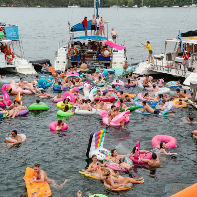 A large group of people enjoying a boat party on a river, with some on boats and others in the water on colorful inflatable floats. In the background, a bridge and city buildings are visible under a cloudy sky. The atmosphere appears lively and fun, reminiscent of events by The Yacht Social Club Sydney Boat Hire.