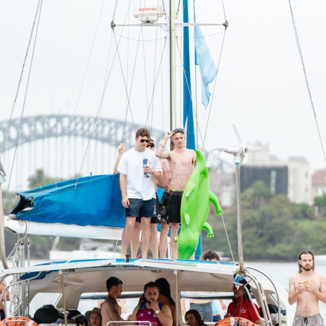 A group of people enjoy a day on a sailboat from The Yacht Social Club Sydney Boat Hire. Two men stand on top of the boat—one holding a green inflatable alligator, the other with a drink. Others relax in different spots, with a famous bridge visible in the background.