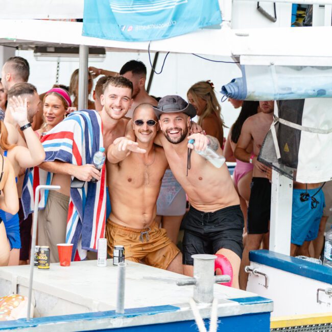 A group of people enjoying a lively boat party hosted by The Yacht Social Club. Three men in swimsuits and sunglasses pose for a cheerful photo, one holding a microphone, another holding a drink. Other guests in bikinis and swim trunks are visible in the background, mingling and having fun on Sydney Harbour.