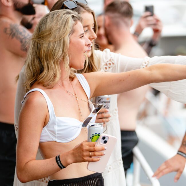 A group of people enjoying a lively and laid-back party on a boat. Several individuals are holding drinks, and a blonde woman in the foreground is pointing while smiling. Most attendees are casually dressed in swimsuits and summer attire, embodying the spirit of Boat Parties Sydney The Yacht Social Club.