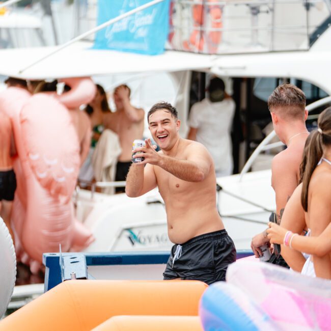 A man in swim trunks stands on a boat holding a drink and smiling at the camera. Behind him are other people in swimsuits, colorful inflatable pool floats, and a yacht. The scene suggests a lively and fun atmosphere with The Yacht Social Club Sydney Boat Hire hosting an exciting party on the water.