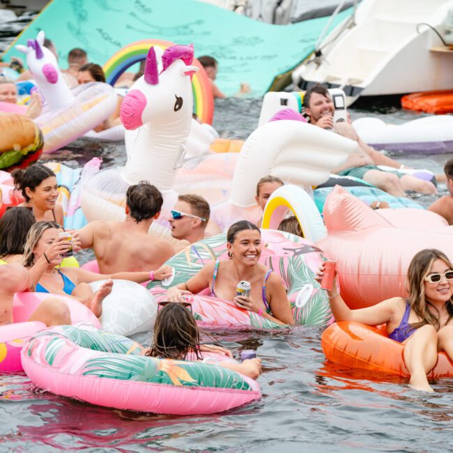 A large group of people are enjoying themselves in the water, floating on various inflatable pool toys, including a unicorn and flamingo. Everyone appears to be having fun under the sun near a docked boat from The Yacht Social Club Sydney Boat Hire, adding to the festive and lively atmosphere.