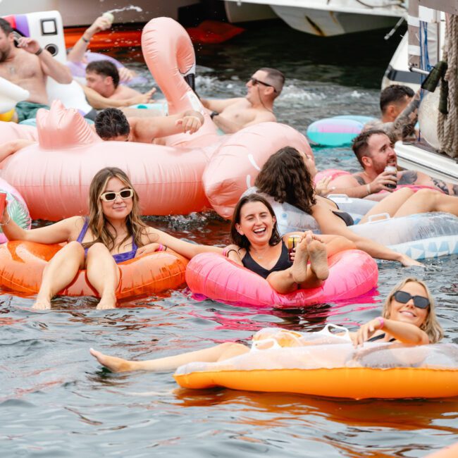 People enjoying a sunny day on the water, lounging on colorful inflatable floats. Some are holding drinks and smiling, while others swim nearby. The background features a boat with more people relaxing and socializing at The Yacht Social Club Sydney Boat Hire. The atmosphere is lively and festive.